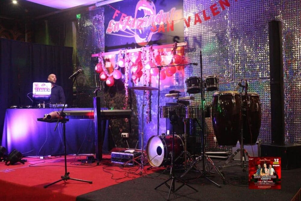 Escenario montado con congas, batería, teclado y cabina de DJ ante un fondo de lentejuelas
